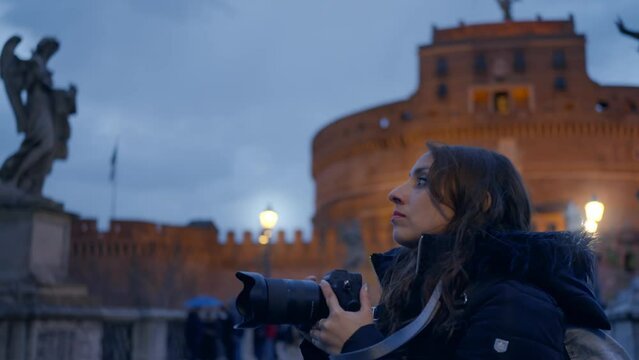 Young Latina woman in warm jacket taking photos on professional photo camera, shooting architecture of Ponte Sant Angelo bridge with lanterns in evening, Rome, Italy