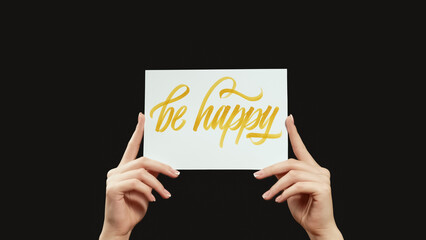 Be happy motivation slogan. Positive attitude. Female hands showing supportive banner with encouraging text isolated on dark black copy space background.