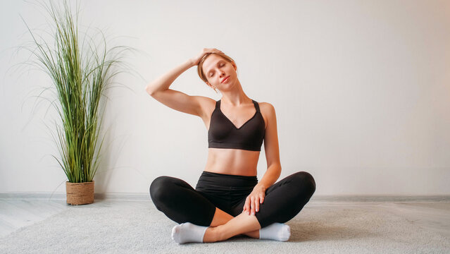 Home yoga. Morning fitness. Flexibility training. Relaxed athletic woman stretching neck sitting cross-legged at light studio interior empty space background.