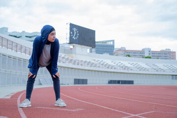 Fototapeta premium A young asian muslim woman wearing a black hijab is doing exercise and resting tired at the outdoor stadium in the morning. Modern Muslim woman concept, Muslim woman sport concept, Islam