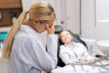 Medium shot of tired stressed female pediatric dentist frustrated with hand on head during appointment of laughing child girl at dental clinic. Concept of children teeth treatment, pediatric checkup.