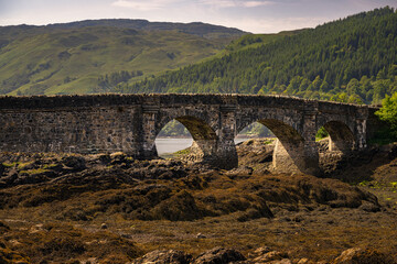 023-06-13 A STONE BRIDGE WITH THREE ARCHES WATER AND TREE LINED HILL IN THE BACKGROUND AND A DRY LAKE BED IN THE FOREGROUND NEAR THE DORINE CASTLE IN SCOTLAND