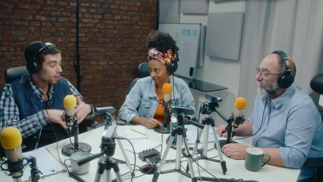 Young blogger in headphones sitting at table with microphones and cameras on tripods, giving an interview or recording podcast with young black woman and senior man in audio studio