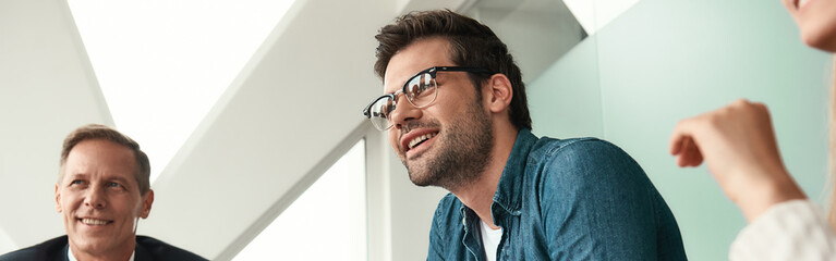 Obraz premium Happy to work here Young and handsome man in eyeglasses discussing something with colleagues and smiling while sitting in the modern office