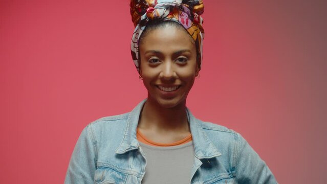 Beautiful young African American woman in casual outfit and head tie standing against a red background and smiling on camera. Studio shot, video portrait