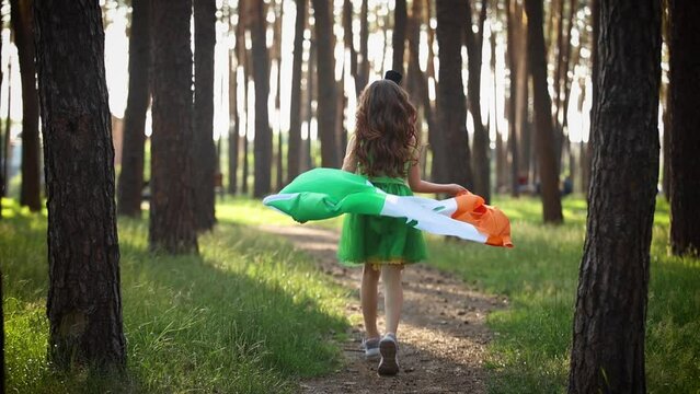 Little girl celebrates St Patrick's Day. Cute 9 year old child in a green pixie costume running through a forest with an Irish flag in slow motion