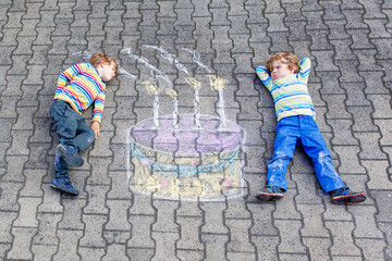 Two happy little kids having fun with big birthday cake picture drawing with colorful chalks....