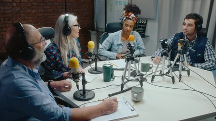 Senior podcast co-hosts recording an episode with young colleagues, sitting all together at table with mics and web cameras on tripods, having discussion in audio studio