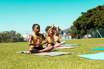 Focused diverse schoolgirls practicing yoga and meditating at stadium on sunny day