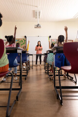 Diverse female teacher with tablet and elementary schoolchildren raising hands in class