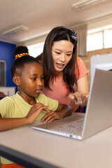 Fototapeta na wymiar Diverse female teacher and schoolgirl using laptop in elementary school class