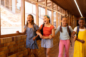 Diverse happy schoolgirls with bags walking and embracing in elementary school corridor