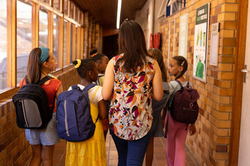 Diverse female teacher and schoolgirls with bags walking in elementary school corridor