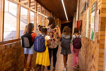 Diverse female teacher and schoolgirls with bags walking in elementary school corridor