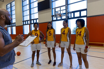 Focused biracial female basketball players with male coach discussing game plan on clipboard at gym
