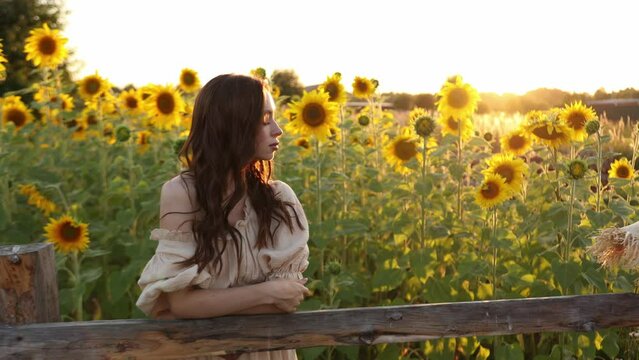 Pretty girl stands among yellow blooming sunflowers. Young woman going through field enjoying freedom and beautiful summer environment. Scenic nature landscape at background. 