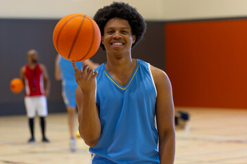 Fototapeta premium Portrait of happy biracial male basketball player doing trick with basketball over teammates at gym
