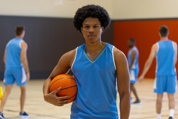 Portrait of biracial male basketball player wearing blue sports clothes over teammates at gym