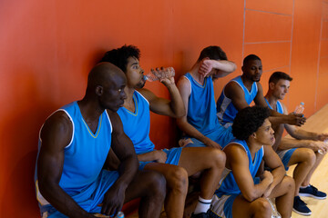 Exhausted diverse male basketball players sitting on bench and drinking water at gym