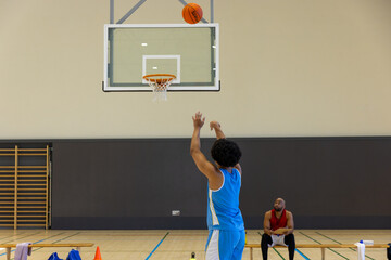 Biracial male basketball player shooting basketball with coach on bench at gym