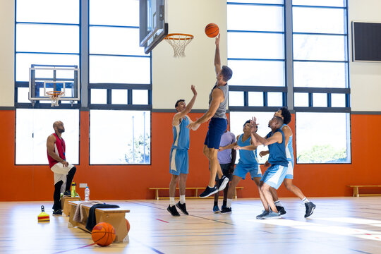Diverse male basketball players wearing blue sports clothes and playing basketball with coach at gym
