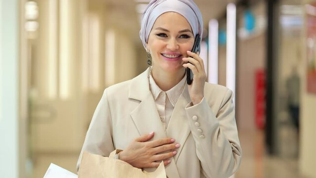 Muslim businesswoman in silver hijab talks with boyfriend on smartphone. Woman carries shopping bags from clothes department in shopping mall