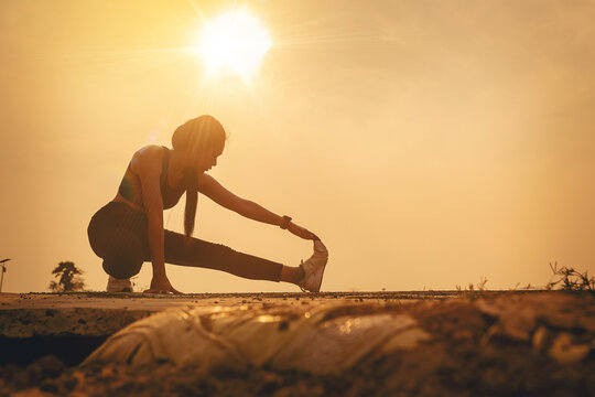 Silhouette of Woman warming up, stretching her muscle at the road track outdoor with sunset background. Fit runner workout and warming up
