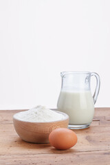 glass jug with milk and wooden bowl with white flour and egg, on wooden rustic table