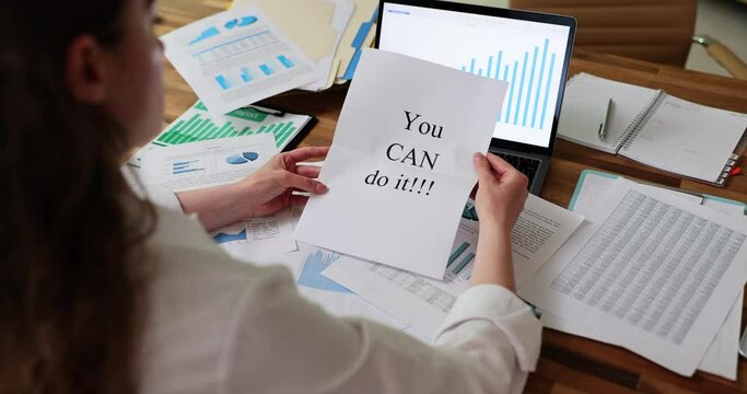 Business lady unfolds piece of paper with motivational phrase You Can Do It sitting at wooden table littered with sheets of papers with statistical data. Employee motivation