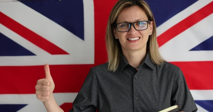 Young woman in glasses shows thumb-up gesture to respect for learning languages. Teacher of English stands at flag of UK encouraging students to study