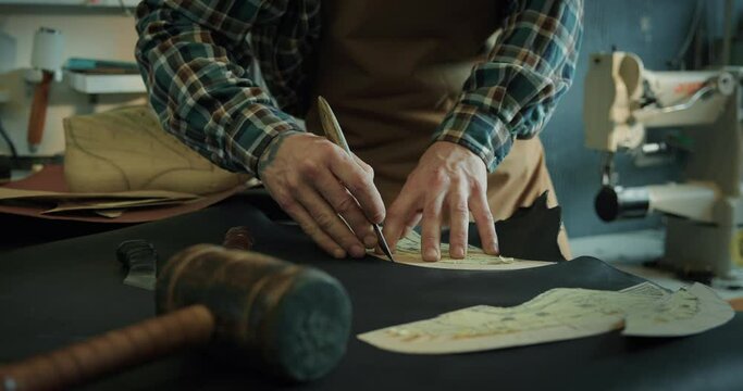 Young male bootmaker cutting out leather pieces on table at the workshop.