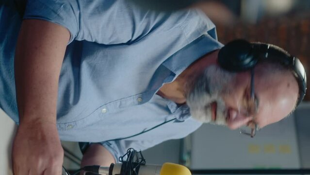 Cheerful senior blogger in headphones sitting by mic and interviewing artist while recording audio podcast in studio. Over-the-shoulder shot, vertical clip