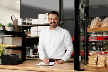 Business owner in his cafe. Man writing notes on clipboard near showcases with pastries
