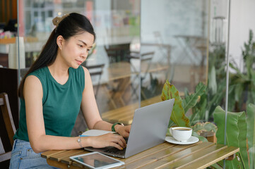 woman working freelance Young Asian woman sitting at work writing articles at wooden table at cafe with laptop and tablet watching webinar writing notes.