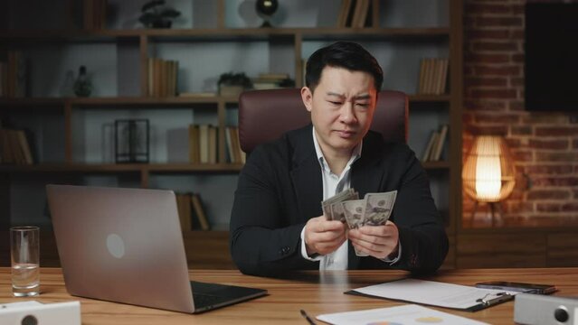 Middle-aged asian office employee counting monthly salary at long wooden table with portable laptop. Man feeling upset and disappointed because of insufficient sum of money.