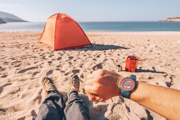 Smart watch on hand, hiker legs with trekking shoes near camping gear and equipment - tent, cooking stove on a sandy beach