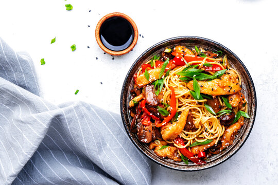 Stir fry noodles with chicken slices, red paprika, mushrooms, chives, soy sauce and sesame seeds in ceramic bowl. Asian cuisine dish. White table background, top view