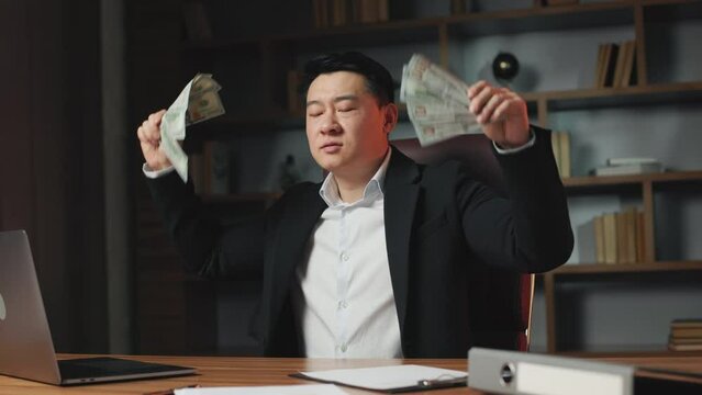 Funny asian millionaire making rhythmical movements with hands full of dollar banknotes. Successful financial director demonstrating enormous company income sitting at desktop in office.