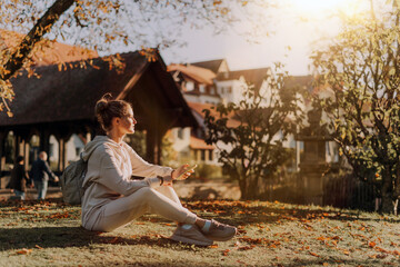Obraz premium Young Fashionable Teenage Girl With Smartphone In Europian Park In Autumn Sitting At Smiling. Trendy Young Woman In Fall In Park Texting. Retouched, Vibrant Colors. Beautiful Blonde Teenage Girl