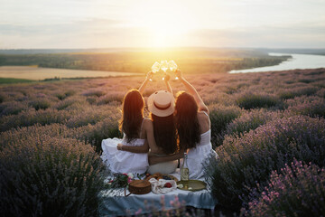 Girlfriends having picnic in the lavender field at sunset. Group of young women sitting on lavender field on summer day. Girlfriends drinking wine on outdoor party.	