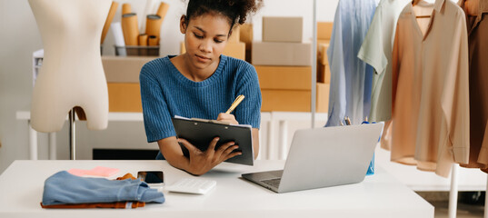 Calm curly brunette dark skinned woman on desk in modern office of fashion designer and holds tablet and smartphone.