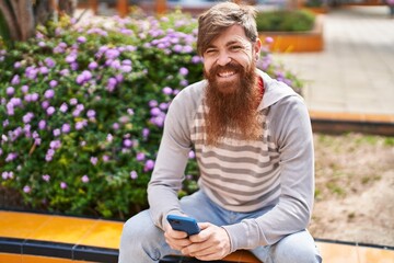 Fototapeta premium Young redhead man using smartphone sitting on bench at park