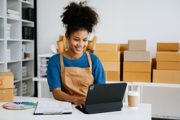 Startup small business SME, Entrepreneur owner African woman using smartphone or tablet taking receive and checking online purchase shopping order to preparing pack product box. in office.