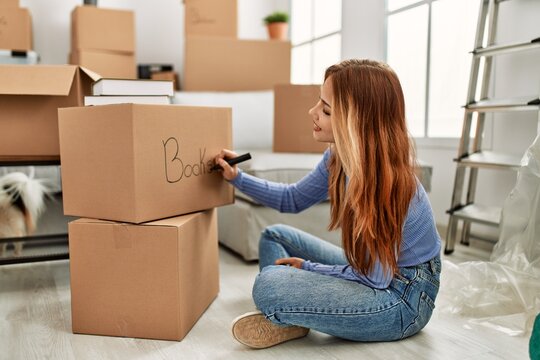 Young caucasian woman smiling confident writing books word on cardboard box at new home
