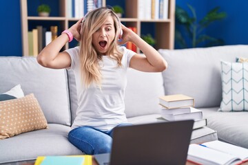 Young blonde woman studying using computer laptop at home crazy and scared with hands on head,...