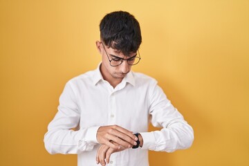 Young hispanic man standing over yellow background checking the time on wrist watch, relaxed and confident