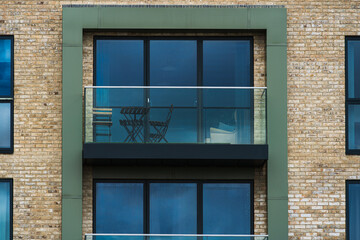 Urban balcony with folding chairs and table, reflected in large apartment windows. Textured brick wall, typical cloudy day in England. Geometric lines of windows and balconies.