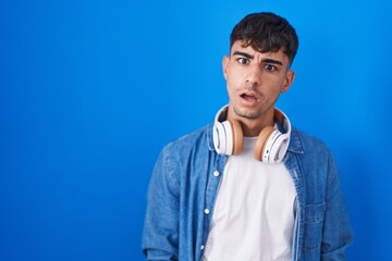 Young hispanic man standing over blue background in shock face, looking skeptical and sarcastic, surprised with open mouth