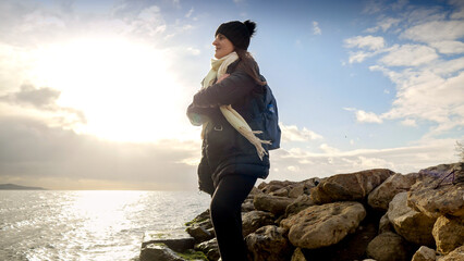 Smiling female tourist wit backpack wearing scarf and hat looking on cold sea waves and sunset