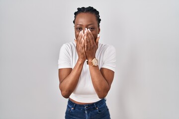 Beautiful black woman standing over isolated background laughing and embarrassed giggle covering...